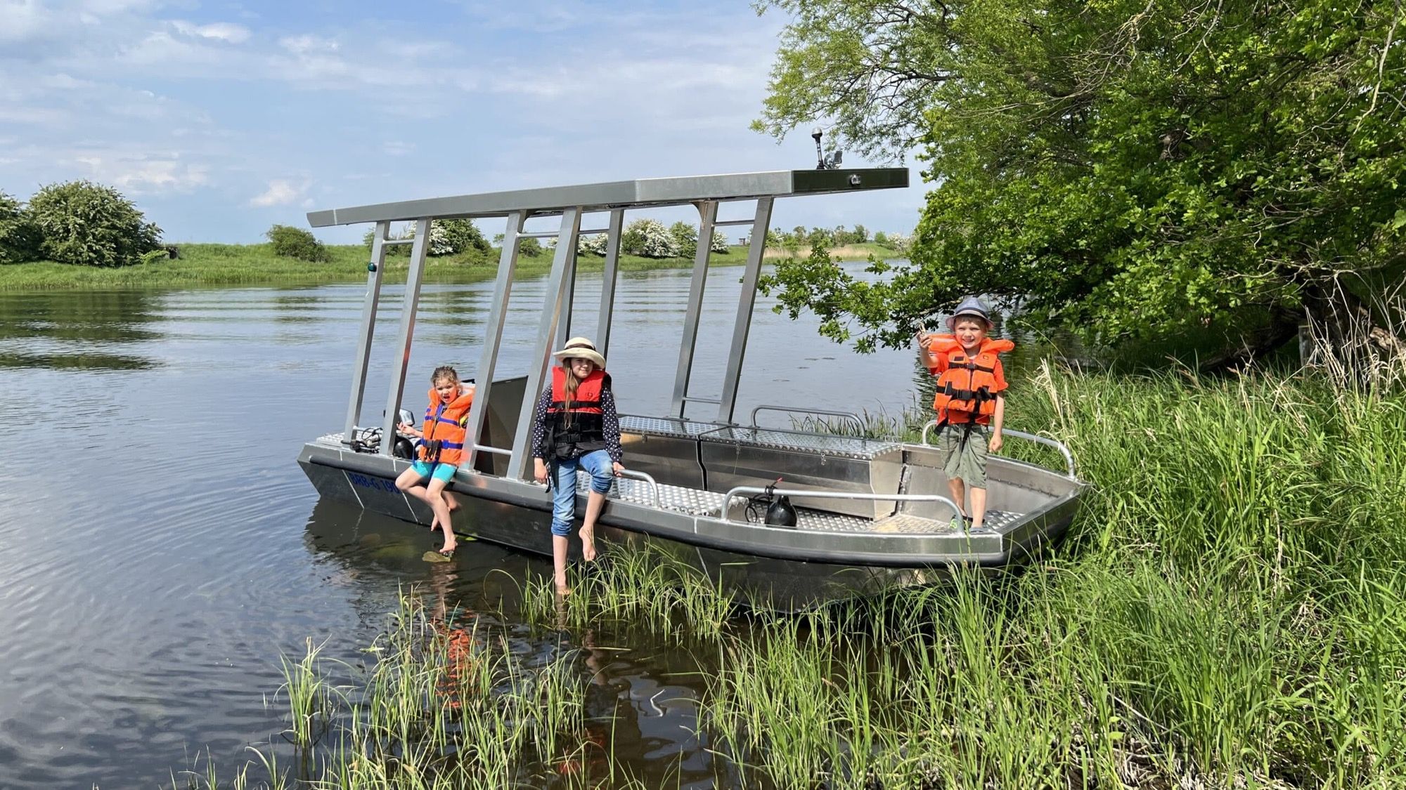 Drei Kinder mit Schwimmwesten an einem flachen Solar-Charterboot am grünen Havelufer bei Havelberg