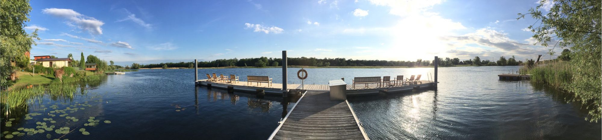 Panorama eines hölzernen Bootsstegs mit Liegen und Bänken, der bei Abendsonne in einen ruhigen See an der Havel ragt